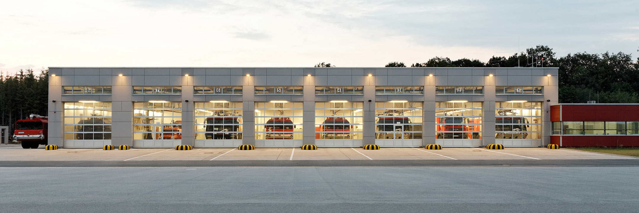 Feuerwache mit verglasten Toren, dahinter mehrere Feuerwehrfahrzeuge in beleuchteter Garage
